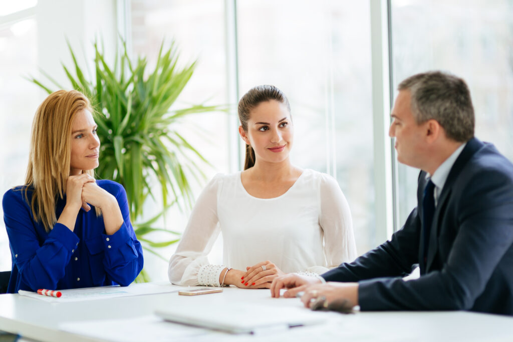 Tres profesionales de negocios, dos mujeres y un hombre, en una reunión formal, discutiendo planes y estrategias futuras. La imagen sugiere un ambiente de asesoramiento y colaboración, donde se analizan datos y se toman decisiones importantes para el crecimiento y el cumplimiento normativo de una empresa.