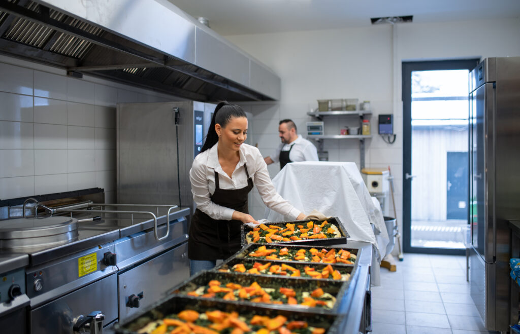 Una chef sonríe mientras prepara bandejas de comida en una cocina industrial, con un colega trabajando en segundo plano. La escena resalta la importancia de los Recursos formativos orientados a hostelería para el manejo eficiente y la preparación de alimentos en grandes volúmenes.