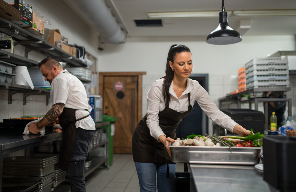 Dos chefs trabajando en una cocina profesional, uno limpiando la encimera de acero inoxidable y otra organizando vegetales frescos. La escena refleja la importancia de los Recursos formativos orientados a hostelería en la gestión de la cocina y el mantenimiento de la higiene alimentaria.