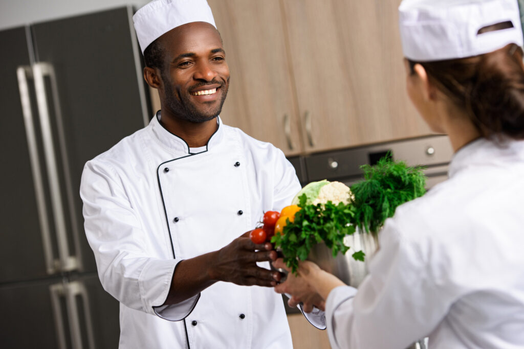 Chef sonriente entregando vegetales frescos a otro chef, simbolizando la formación continua y el desarrollo profesional, aspectos clave de los Derechos y Deberes en Hostelería España.