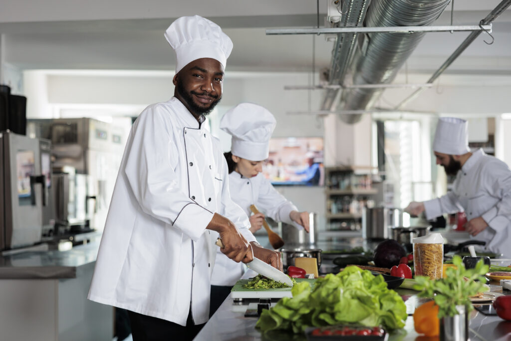 Chef sonriente picando vegetales en una cocina profesional, con otros chefs al fondo, representando la importancia de los Derechos y Deberes en Hostelería España y la seguridad en el sector.